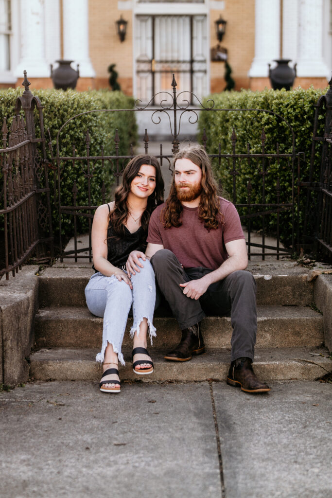 Couple sitting on a stoop in front of a metal fence in front of green hedges for their engagement session